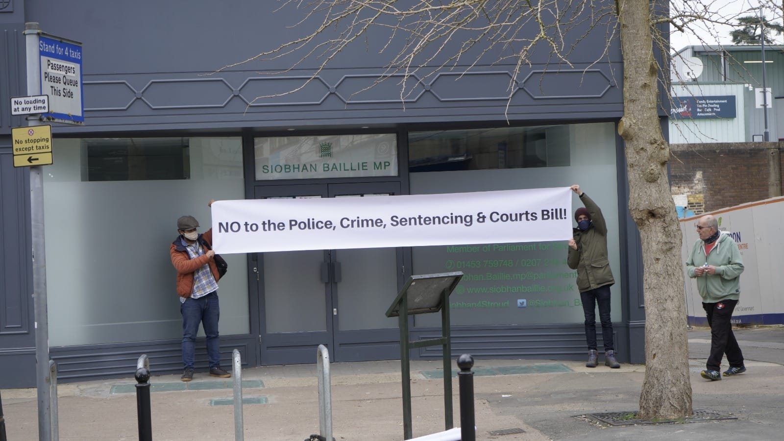 Two people standing outside Stroud MP Siobhan Baillie's office holding up a long white banner with black text that reads "NO to the Police, Crime, Sentencing & Courts Bill!"