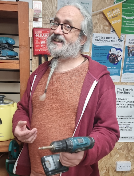 A picture of a smiling grey-haired man with an elaborately knotted beard. He is wearing an orange jumper and a red hoodie. In his left hand he holds a cordless drill.