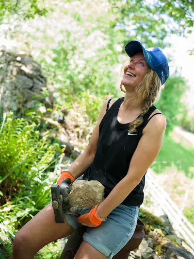 A photograph of Kirsty leaning against a wall, smiling and holding a large stone in her hands