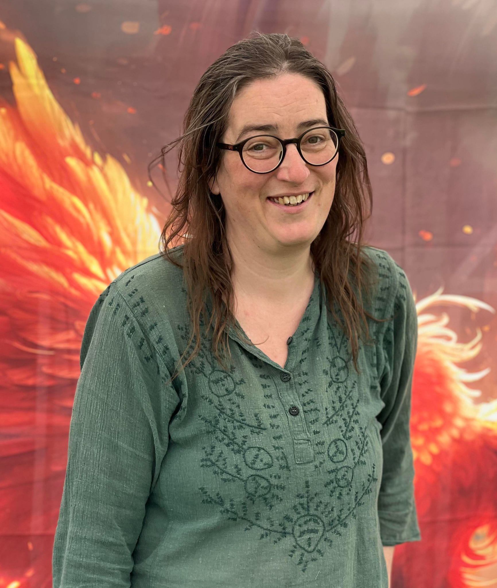Nimue Brown weating a dark green patterned top and standing in front of a bright orange phoenix wing art display. Nimue has long brown hair and is wearing rounded glasses. She is smiling to the camera