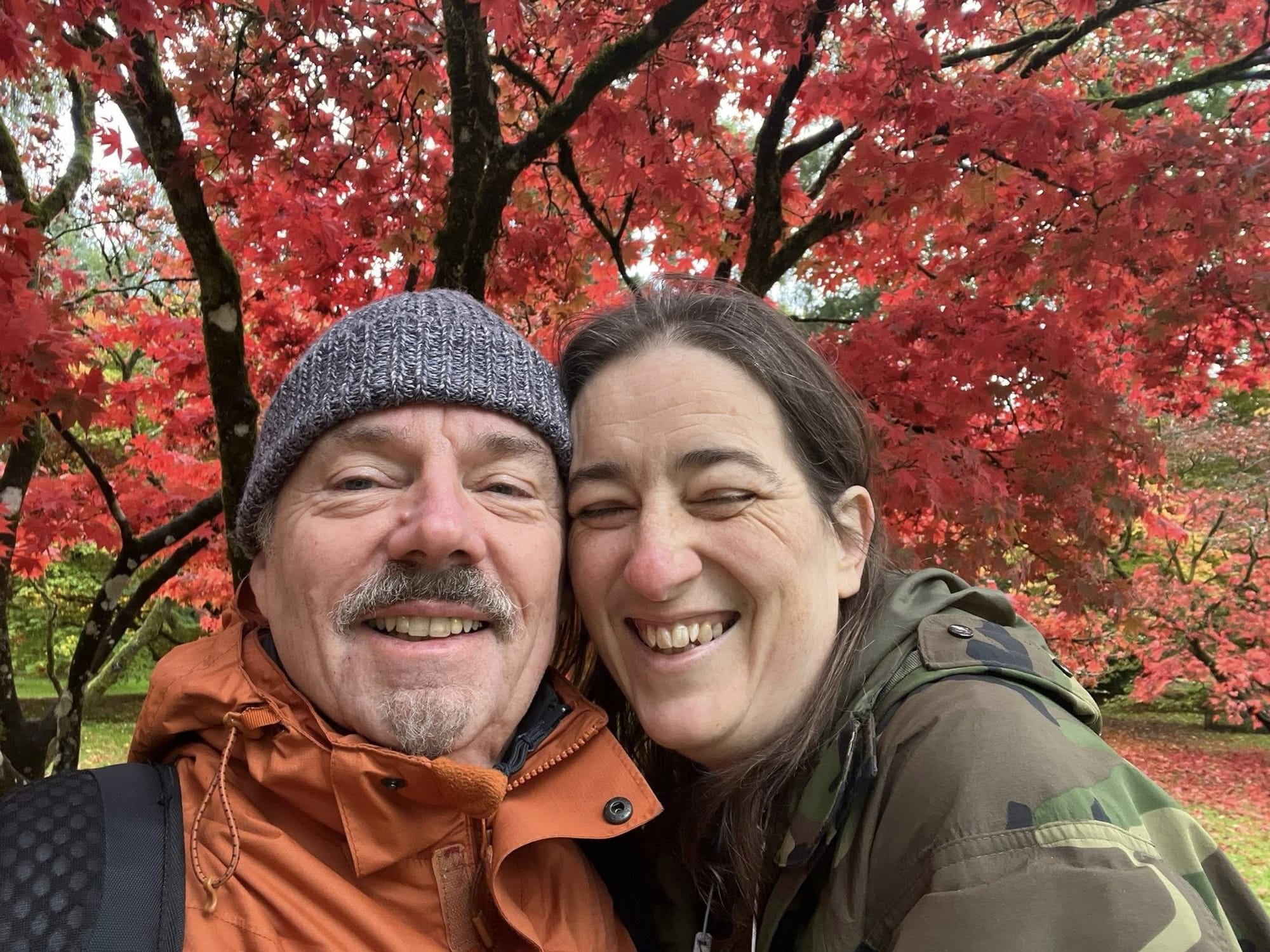 A smiling man with a moustache and a goatee beard, wearing a grey beanie hat and an orange jacket is being embraced by a woman in a camouflage coat. The woman is also smiling and has her eyes closed. They are standing in front of a beautiful red Maple tree on a patch of grass at Westonbirt Arboretum