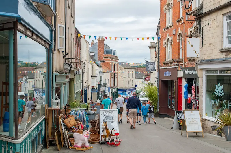 A view down Stroud High Street. A row of shops on either side with 'A' boards, rocking horses, bric-a-brac and other assorted items spilling out onto the pavement that is already steep and uneven