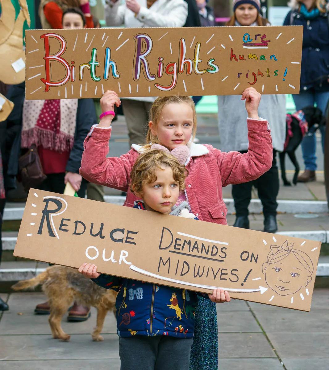 Two young children hold placards: one reads "Birth Rights — Human Rights" and the other "Reduce our demands on midwives"
