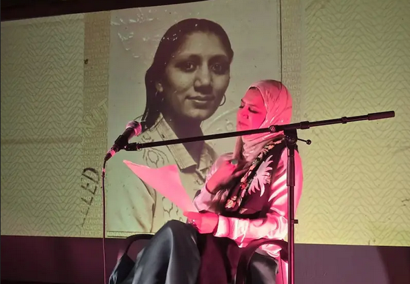 A young woman wearing a hijab sits and reads from a script in front a photograph of another woman
