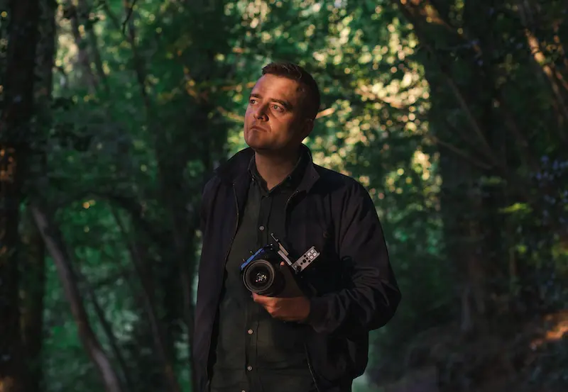 Picture of a man holding a video camera and staring thoughtfully into the distance. The man is in a woodland area.